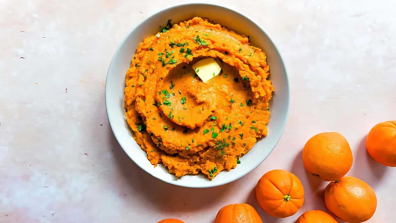 A bowl of mashed sweet potatoes with a pat of butter and chopped herbs on top, surrounded by whole oranges on a light surface.