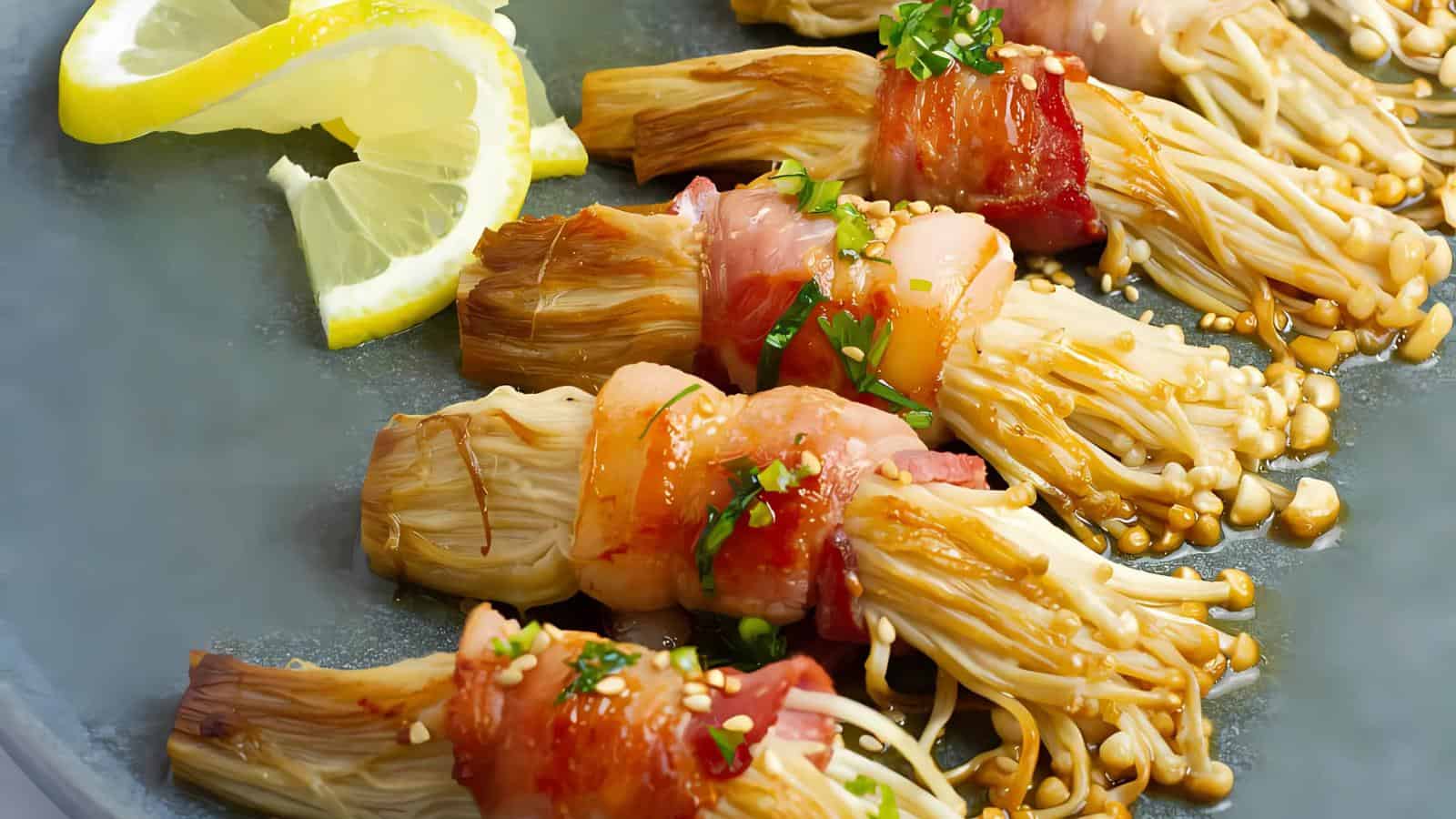 Bacon-wrapped enoki mushrooms garnished with sesame seeds and herbs, served with lemon wedges on a gray plate.