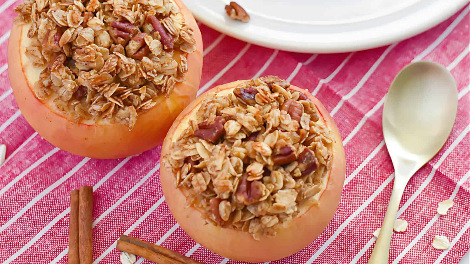 Two baked apples filled with granola and nuts sit on a red striped cloth, next to a spoon and cinnamon stick. A white plate is partially visible in the background.