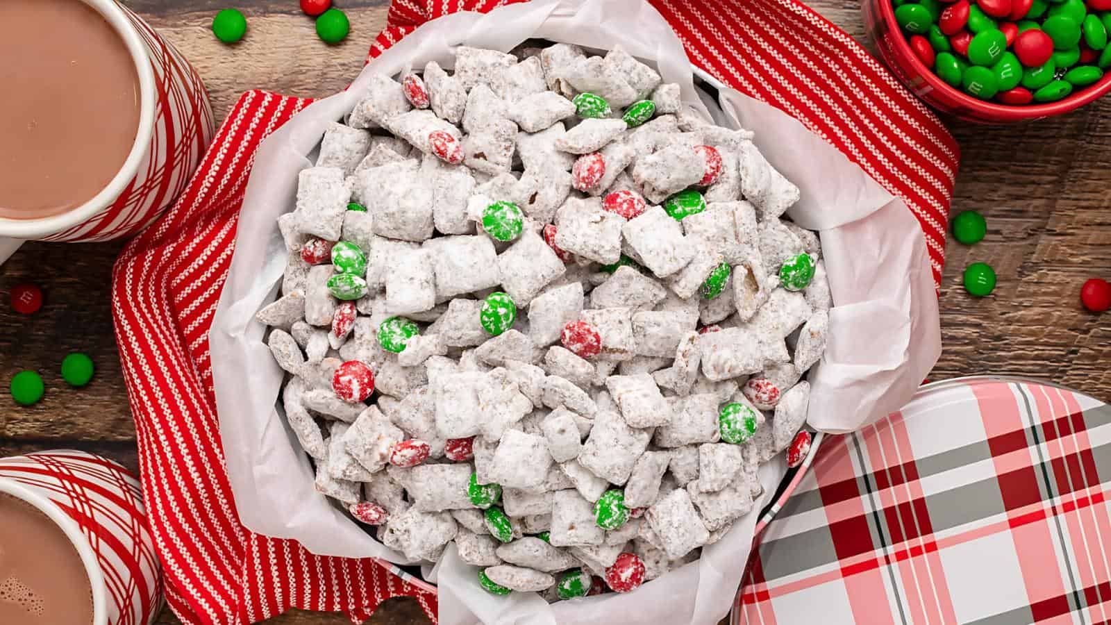 A bowl of powdered sugar-coated snack mix with red and green candies, surrounded by festive decor and mugs of hot chocolate.