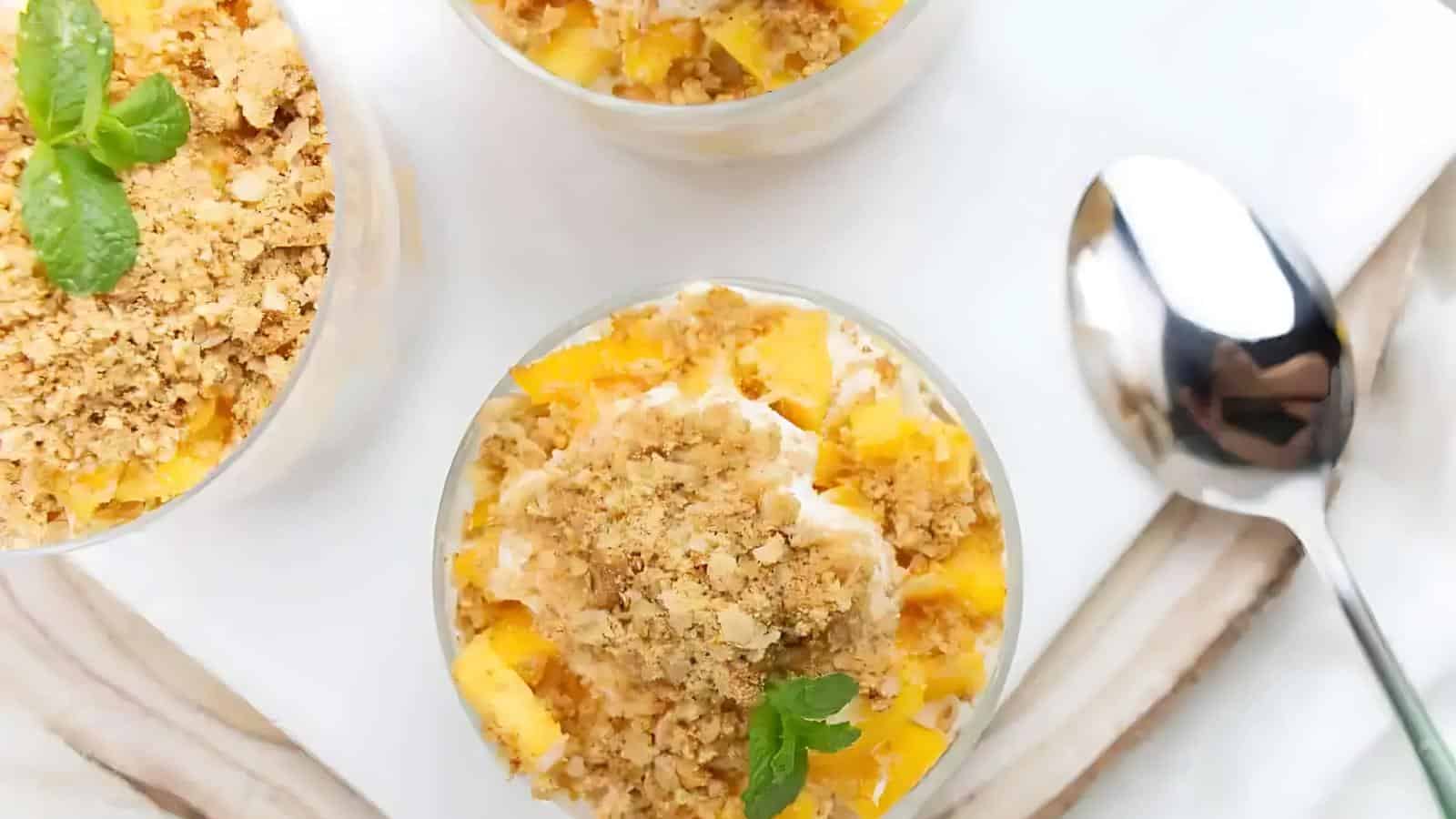 Three dessert cups with crumble topping and mint garnish, placed on a white surface beside a spoon.