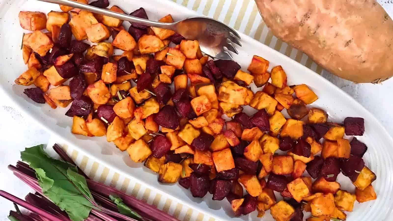 A dish of roasted sweet potatoes and beets on a striped cloth, with a metal serving spoon, a whole sweet potato, and beet greens beside it.