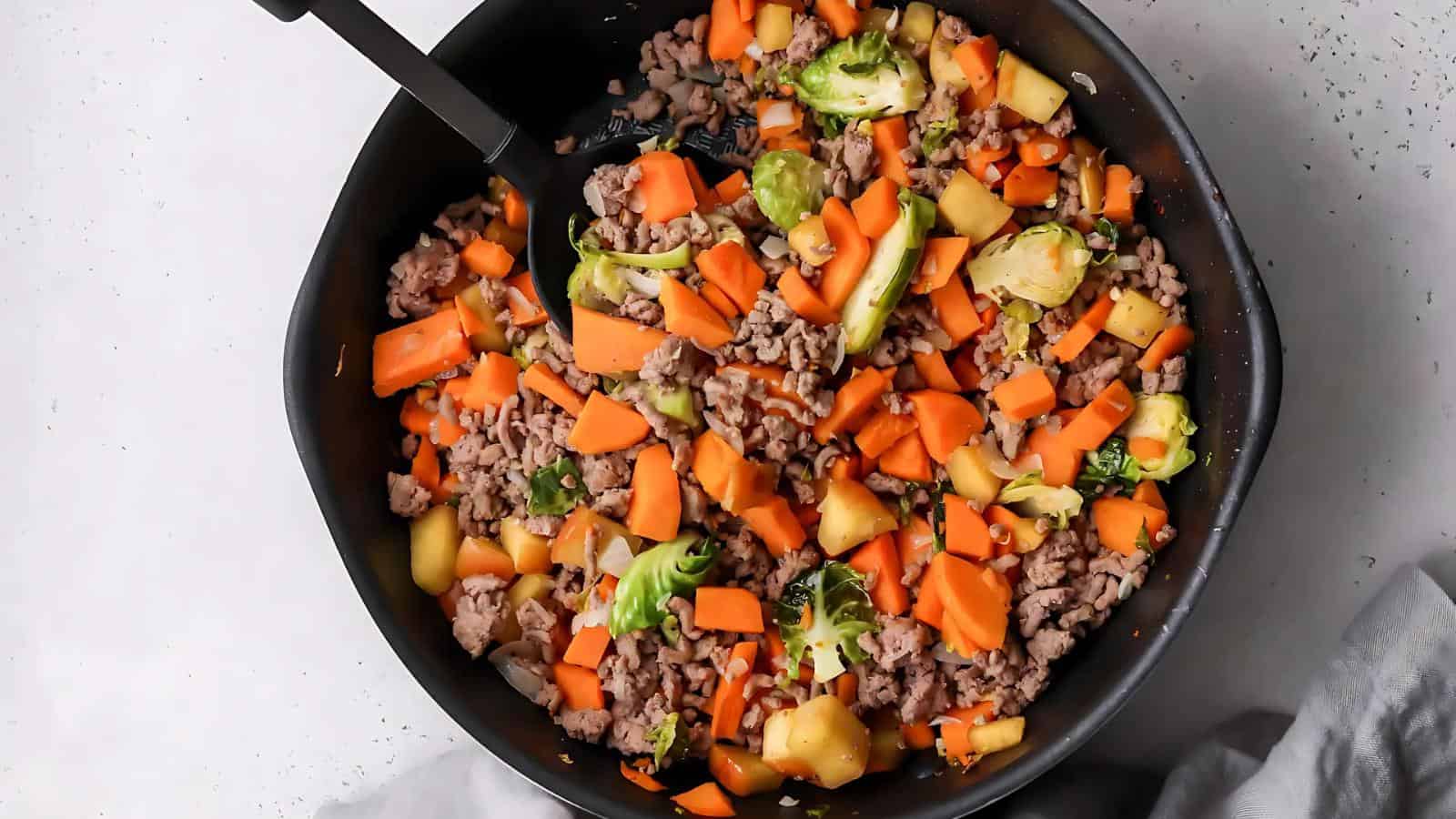 A skillet with ground meat, diced carrots, sliced Brussels sprouts, and chopped pineapple chunks being mixed with a black spatula.
