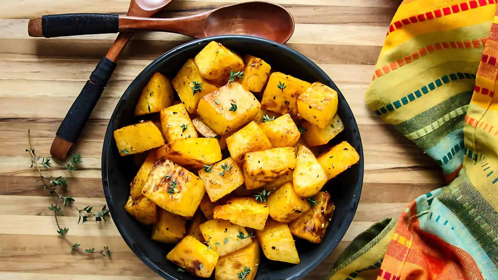 A bowl of roasted cubed butternut squash garnished with herbs on a wooden table, next to a spoon and a colorful striped cloth.