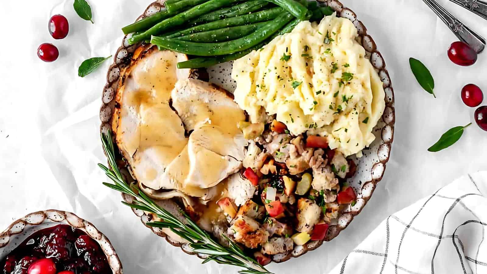 A plate with sliced turkey, green beans, mashed potatoes, stuffing, and a sprig of rosemary, accompanied by a small dish of cranberry sauce.