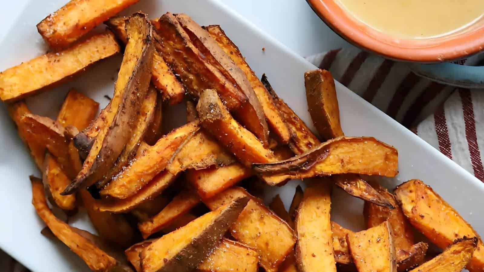 A white plate filled with crispy sweet potato fries alongside a small bowl of dipping sauce.