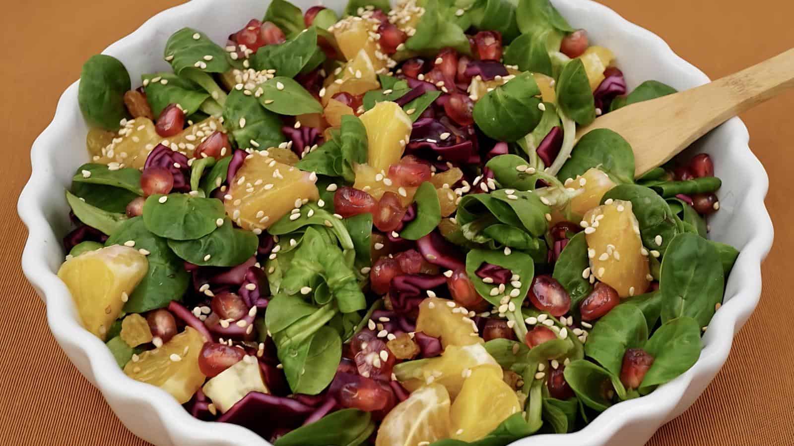 A mixed salad in a white bowl containing spinach, oranges, red cabbage, pomegranate seeds, and sesame seeds, with a wooden spoon on the side.