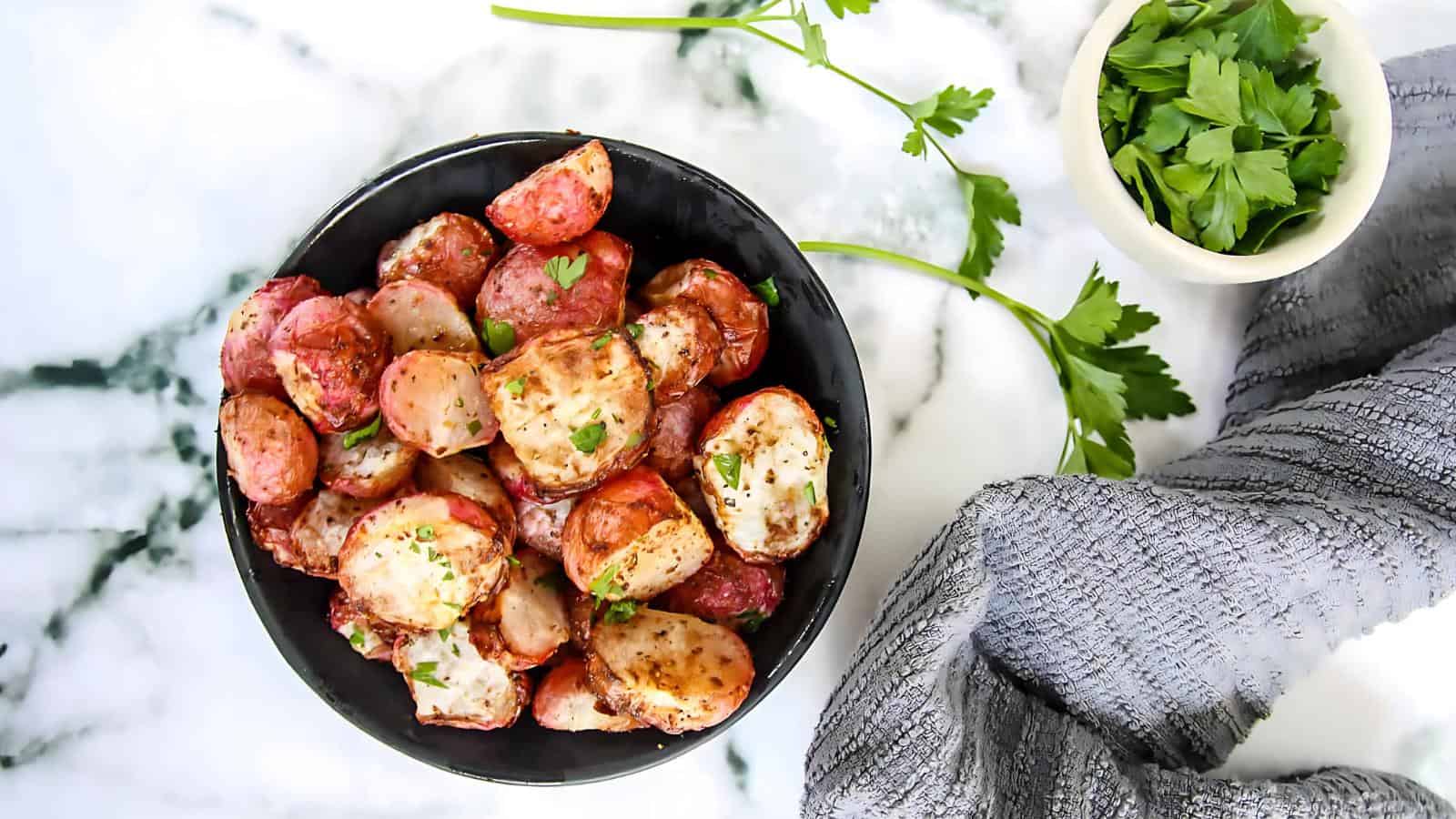 A black bowl of roasted red potatoes garnished with herbs on a marble surface, accompanied by a small dish of fresh parsley and a textured gray cloth.