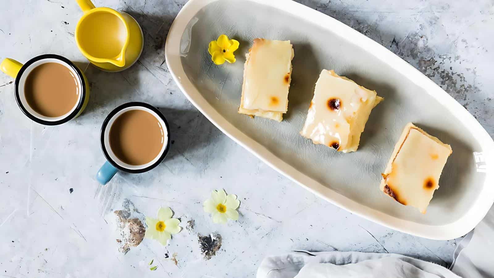 Two cups of coffee next to a tray with three glazed pastries and small yellow flowers on a light-colored surface.