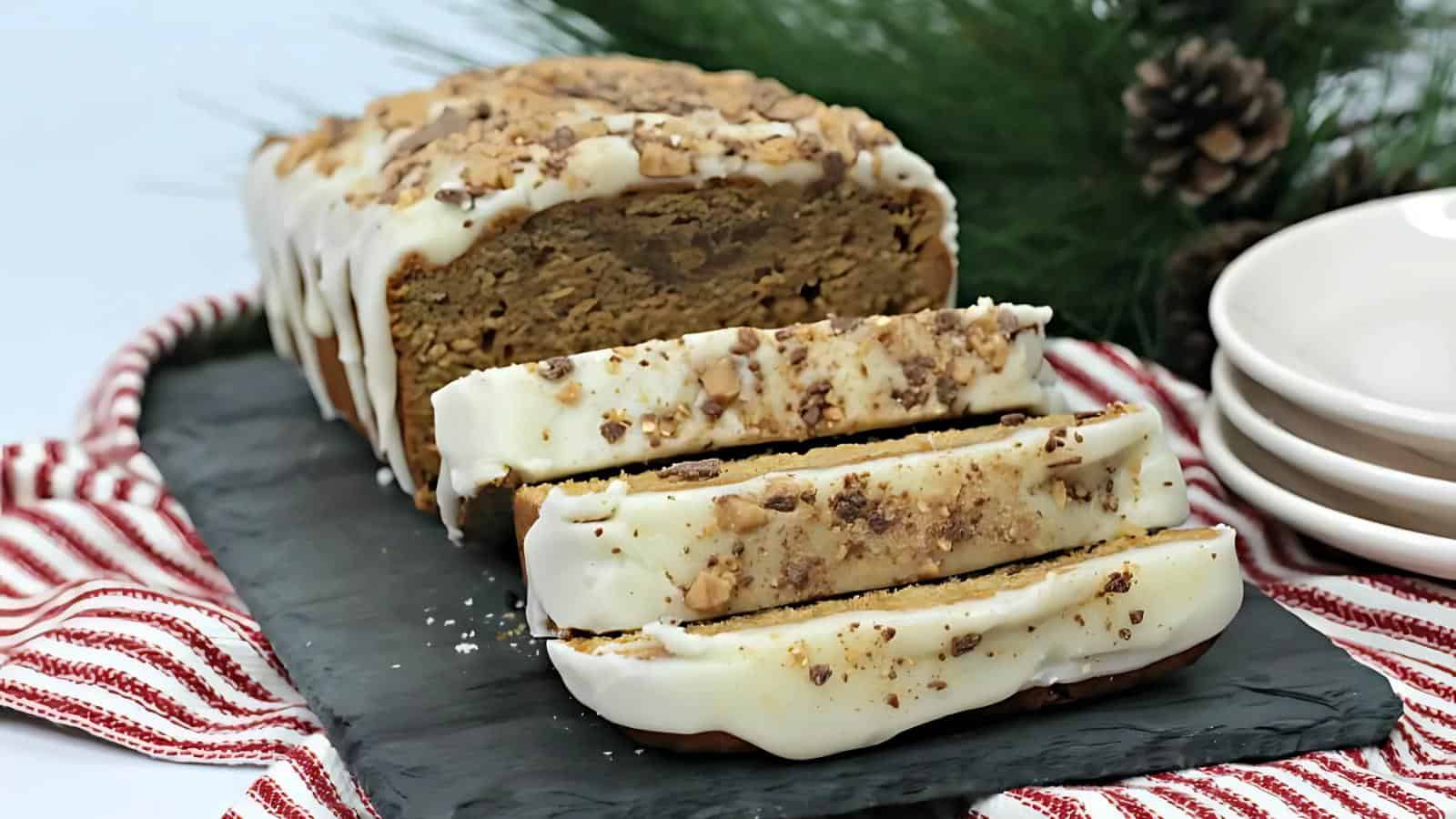 Sliced loaf of bread with white icing and crumble topping on a slate board, next to stacked plates and pine branches.