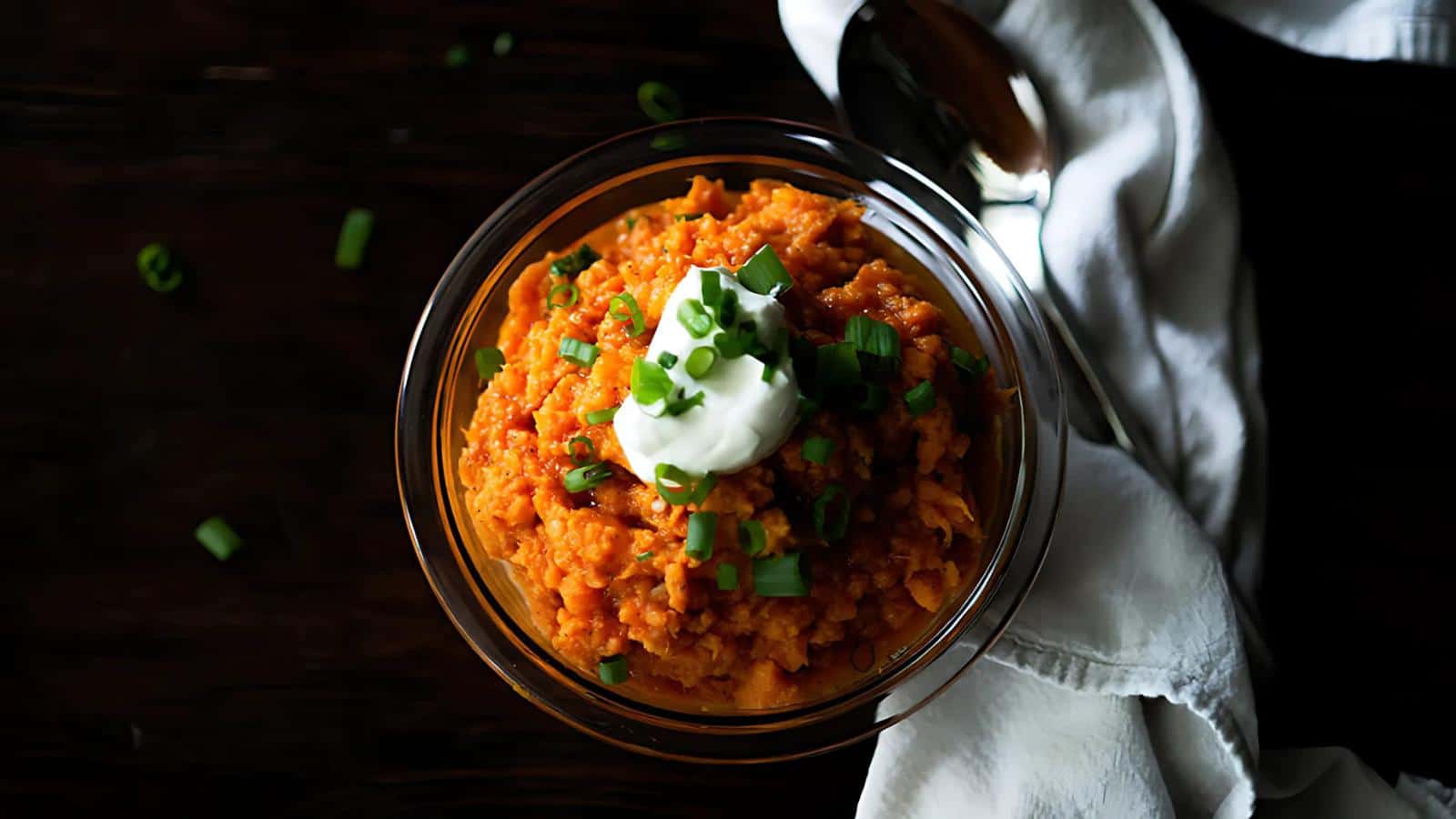A bowl of mashed sweet potatoes topped with sour cream and chopped green onions, accompanied by a spoon and a white cloth on a dark surface.