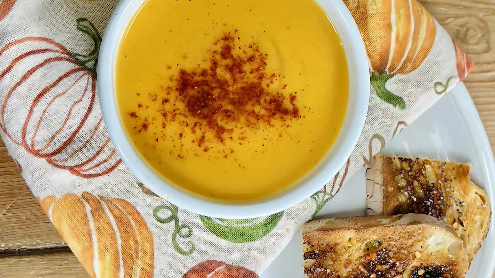 A bowl of bright orange soup with paprika on top, placed on a pumpkin-patterned cloth. Slices of toasted bread with seeds are on the side.