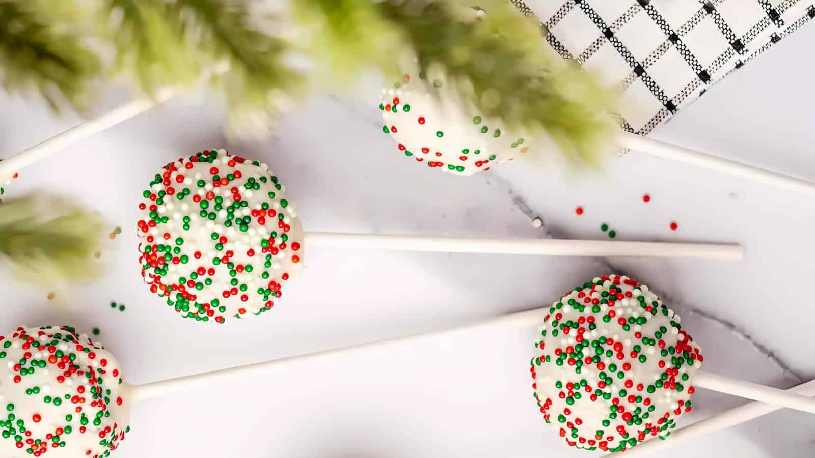 Four white cake pops covered with red and green sprinkles on a white background, next to a black and white checkered cloth.