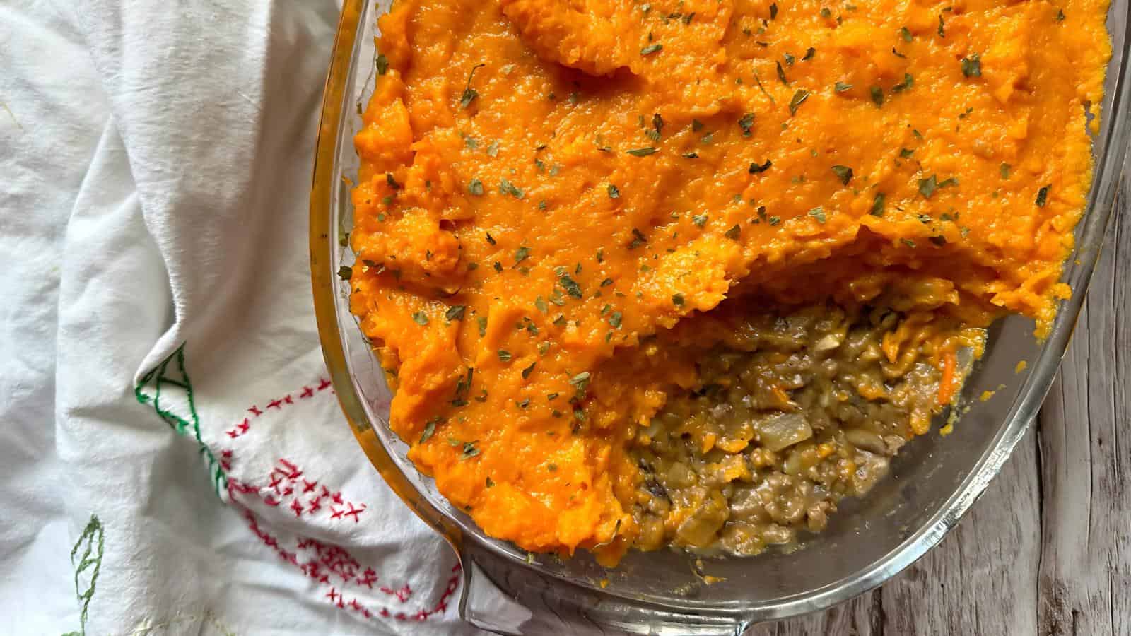 A dish with mashed sweet potatoes on top of a ground meat and vegetable filling, partially served, next to a white embroidered cloth.