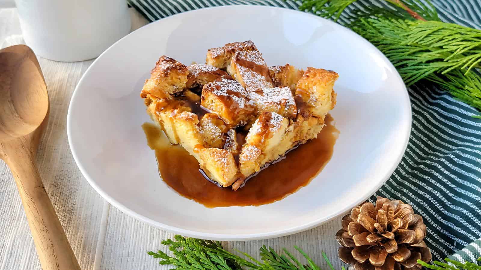 A plate of bread pudding topped with powdered sugar and caramel sauce, surrounded by pine elements and a wooden spoon.