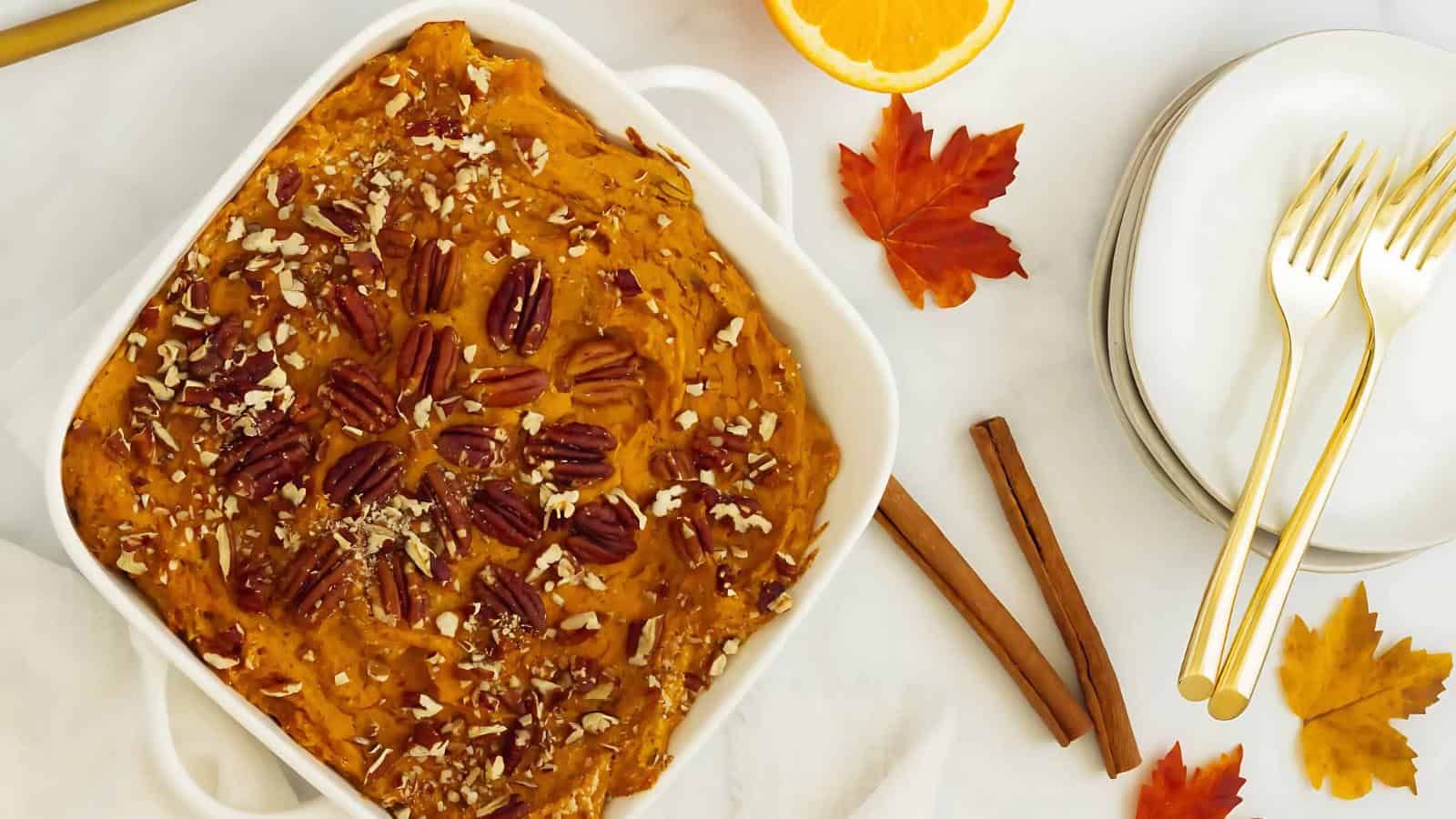 A baked dish of sweet potato casserole topped with pecans in a white square dish, surrounded by autumn leaves, cinnamon sticks, a slice of orange, and stacked plates with forks.