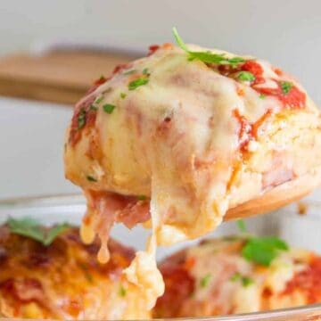 Close-up of a freshly baked lasagna serving being lifted from a glass dish with a wooden spoon. The lasagna is topped with melted cheese and garnished with parsley.