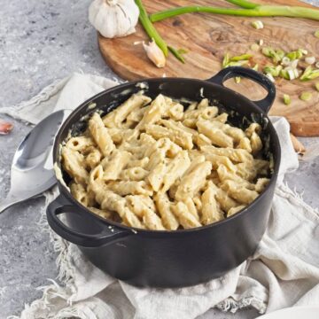 A black pot filled with creamy penne pasta, placed on a white cloth next to a wooden board with chopped green onions and garlic on a gray surface.