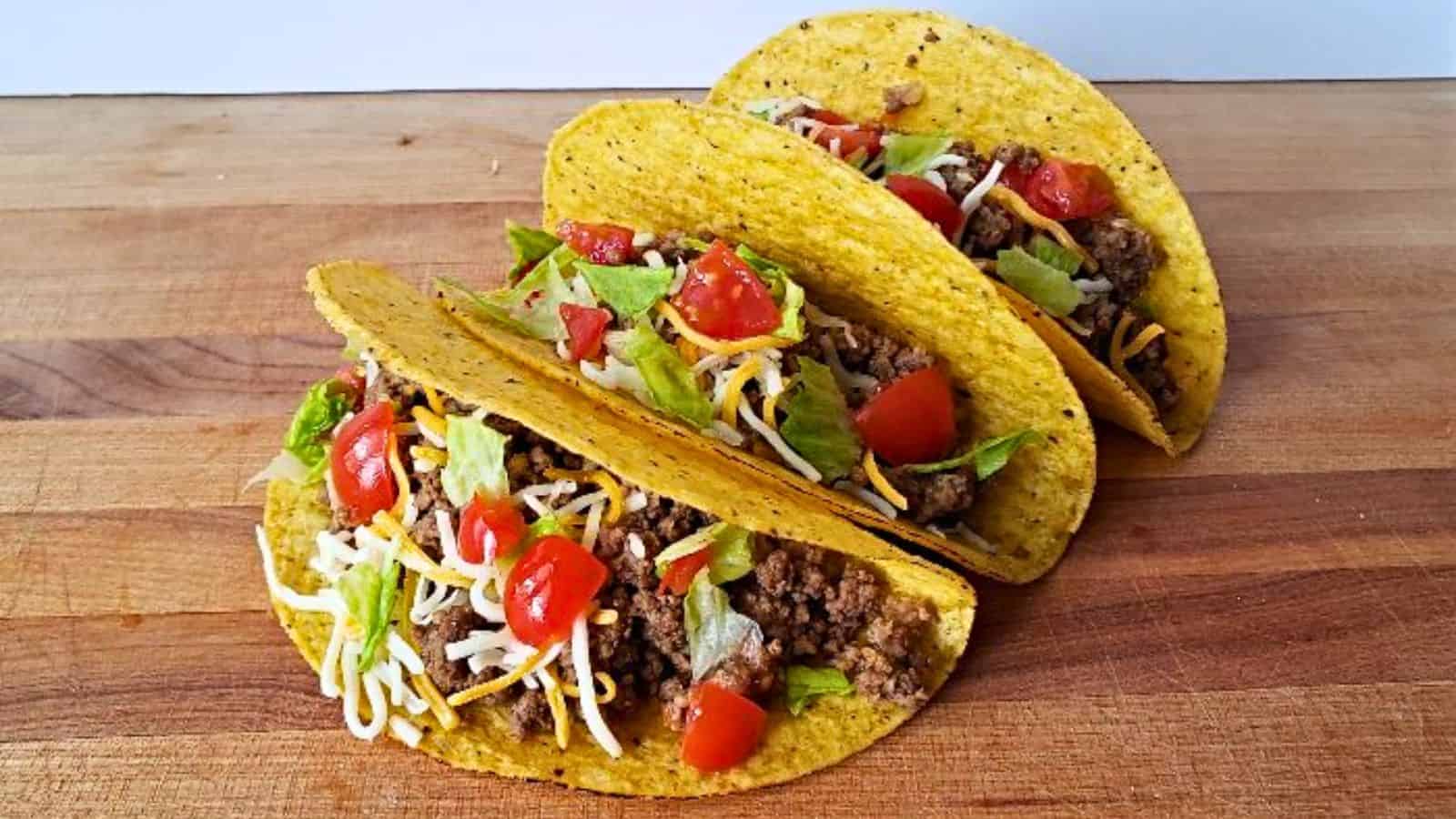 Three hard-shell tacos filled with ground beef, lettuce, diced tomatoes, and shredded cheese are arranged on a wooden surface, making them ideal as easy Mexican inspired meals for the ultimate lazy cooks.
