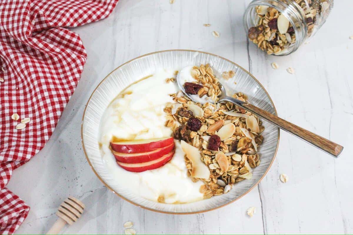 A bowl of yogurt with sliced apples, granola, and a drizzle of honey on a white surface. A spoon rests inside, with a checked cloth and a jar of granola nearby.