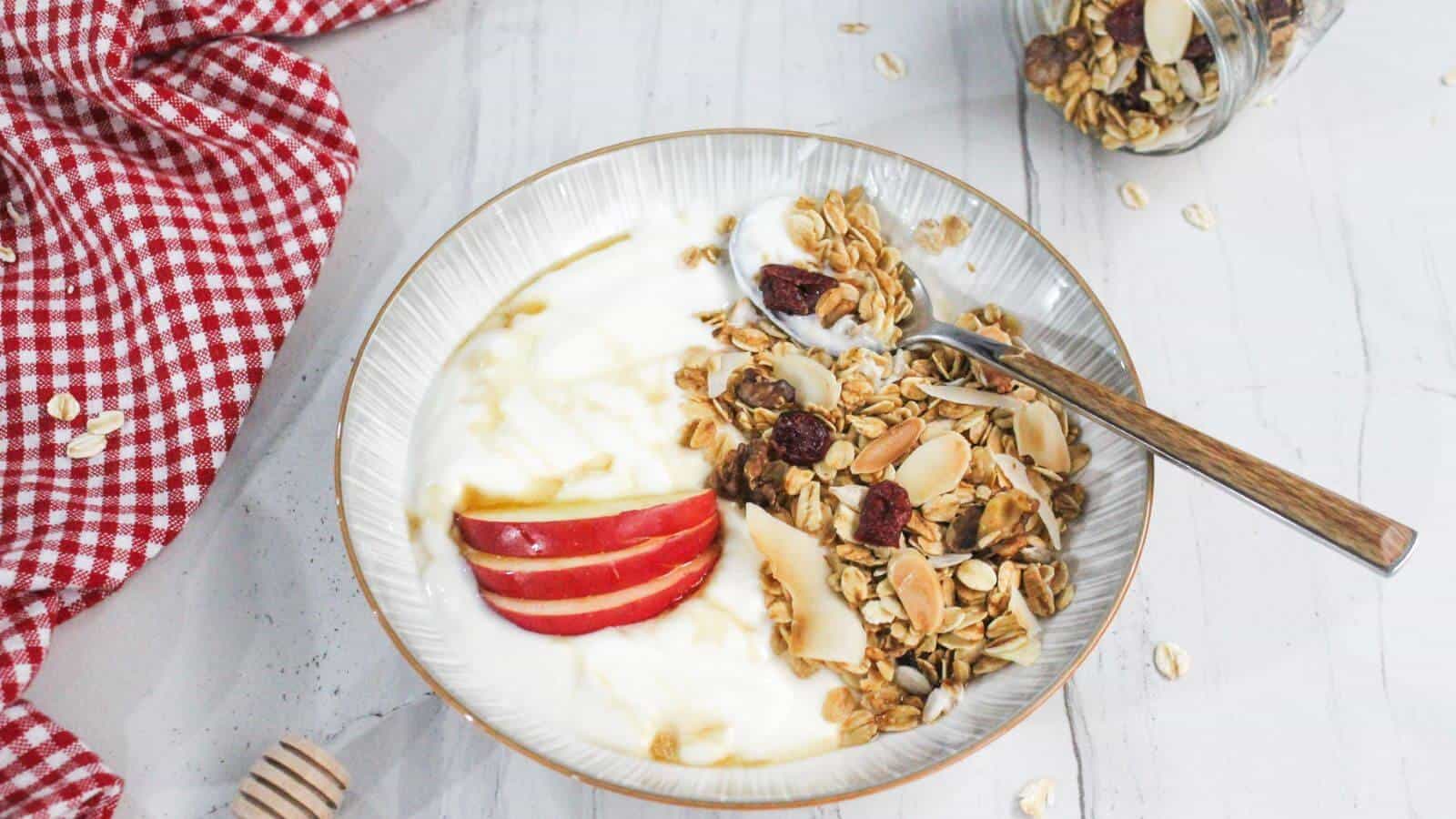 A bowl of yogurt topped with sliced red apples, granola, and dried fruit, with a spoon resting inside. A jar of granola is tipped over nearby on a white surface with a red checkered cloth.