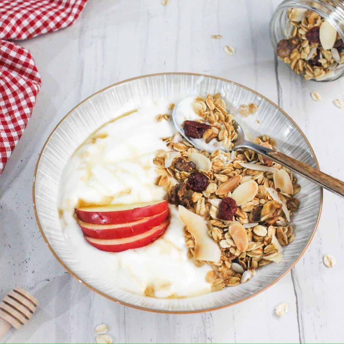 A bowl of yogurt topped with granola, sliced red apples, and a drizzle of honey. A spoon is placed in the bowl. A red checkered cloth is in the background.