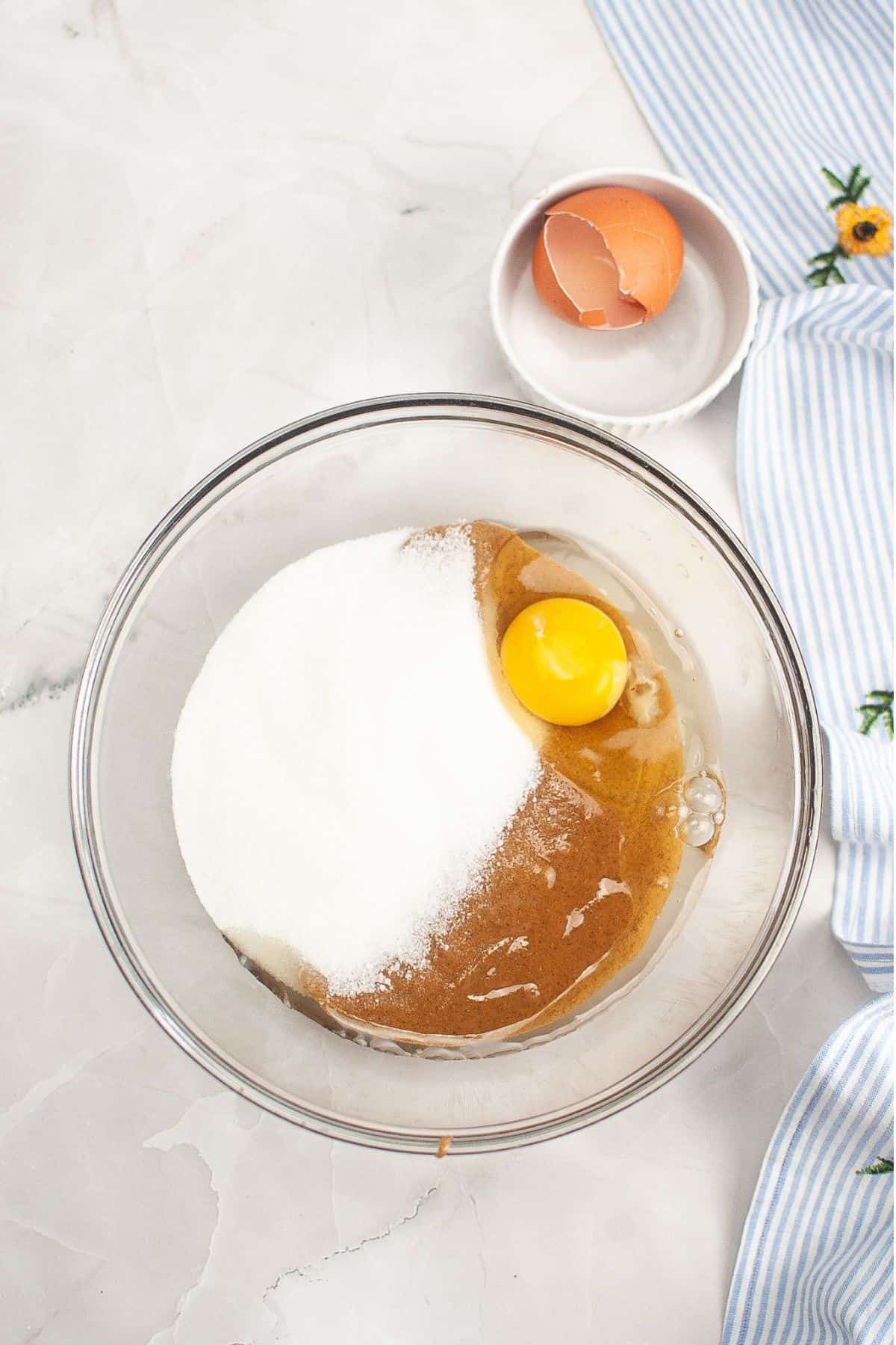 A mixing bowl with sugar, brown sugar, and an egg. An egg shell is on a plate nearby, and part of a striped cloth is visible.