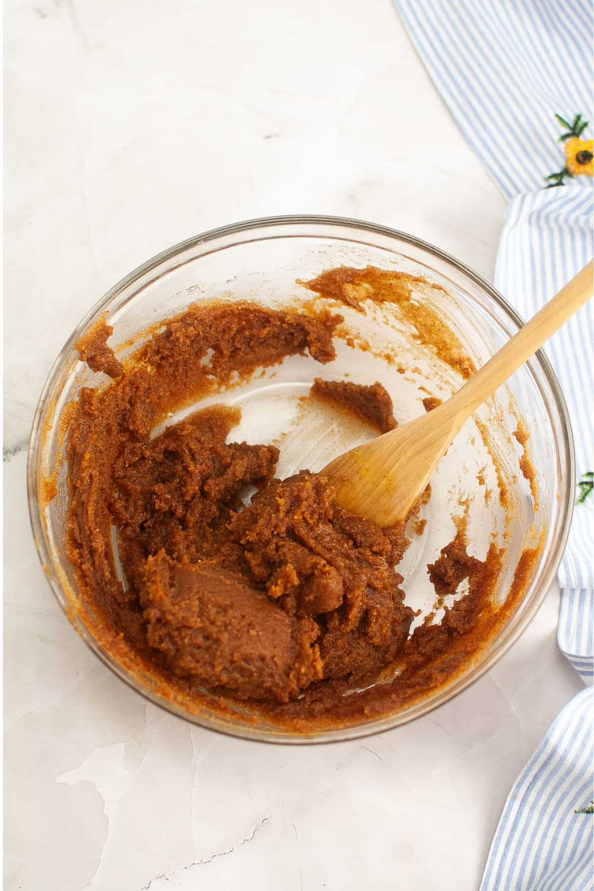 A glass bowl filled with brown cookie dough and a wooden spoon on a white surface near a blue-striped cloth.
