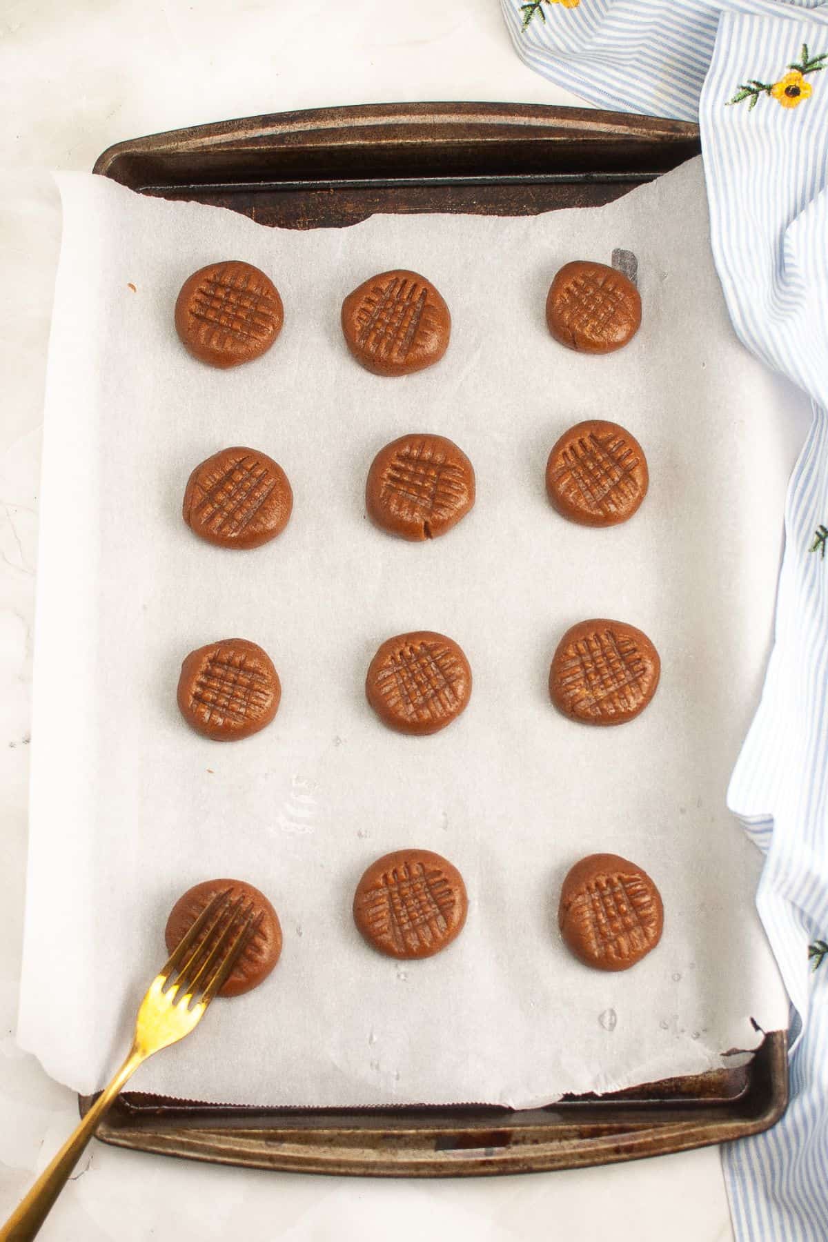 A baking tray with 14 unbaked peanut butter cookies on parchment paper. A fork rests on the tray, showing the crisscross pattern on one cookie. A striped cloth lies beside the tray.