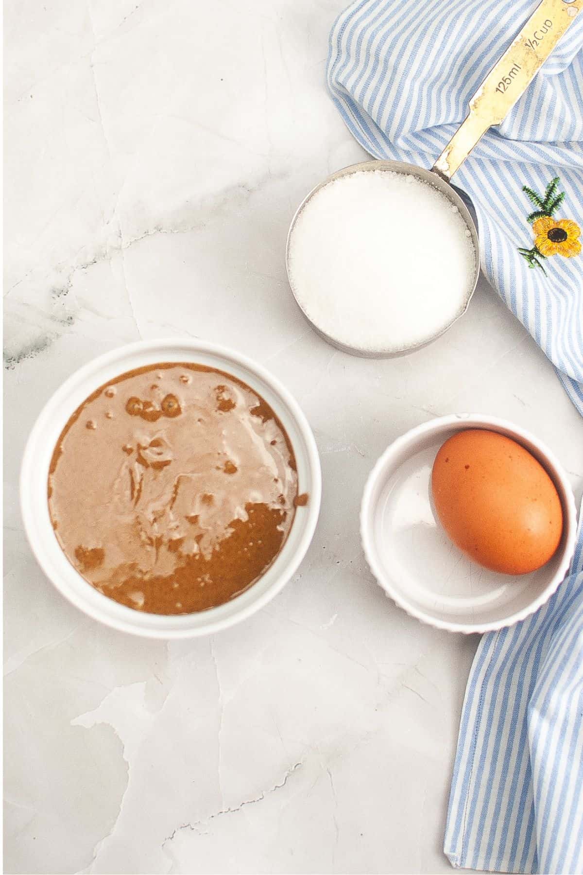 A bowl of brown batter, a whole egg, a cup of sugar, and a striped cloth on a marble surface.