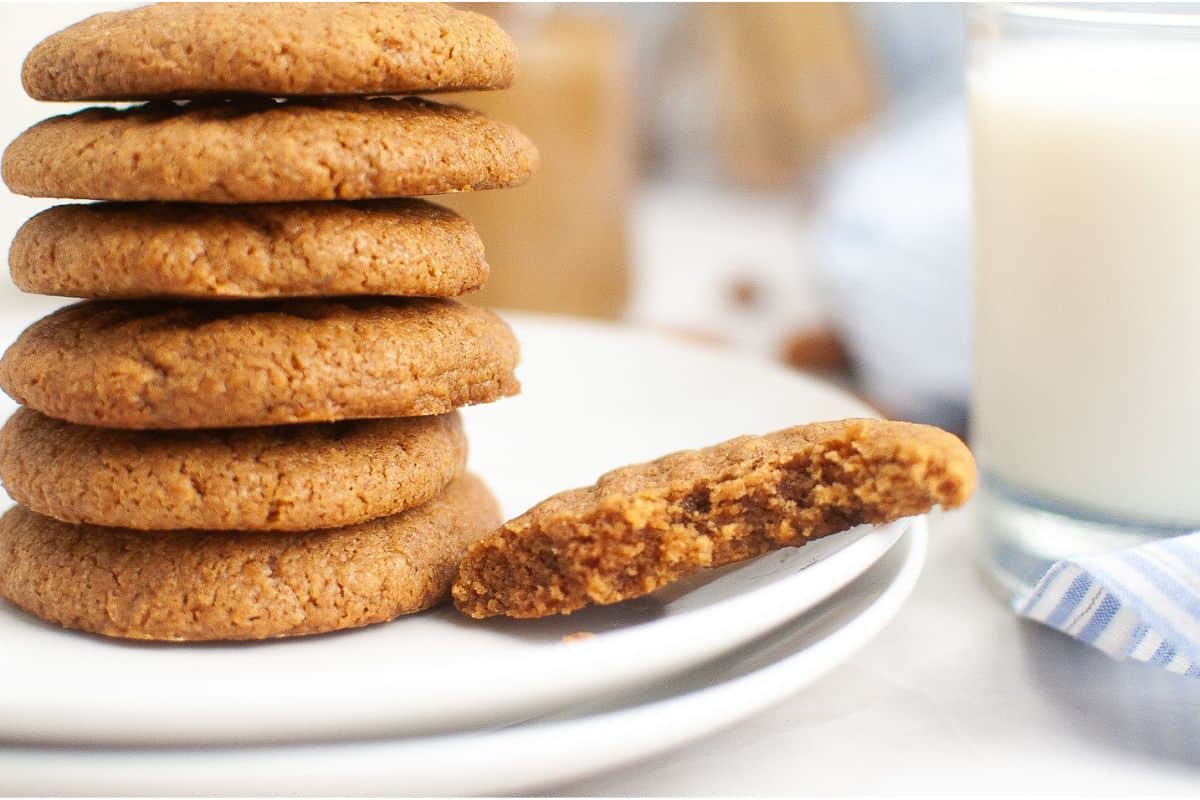 A stack of cookies on a white plate with a partially eaten cookie in front. A glass of milk is in the background.