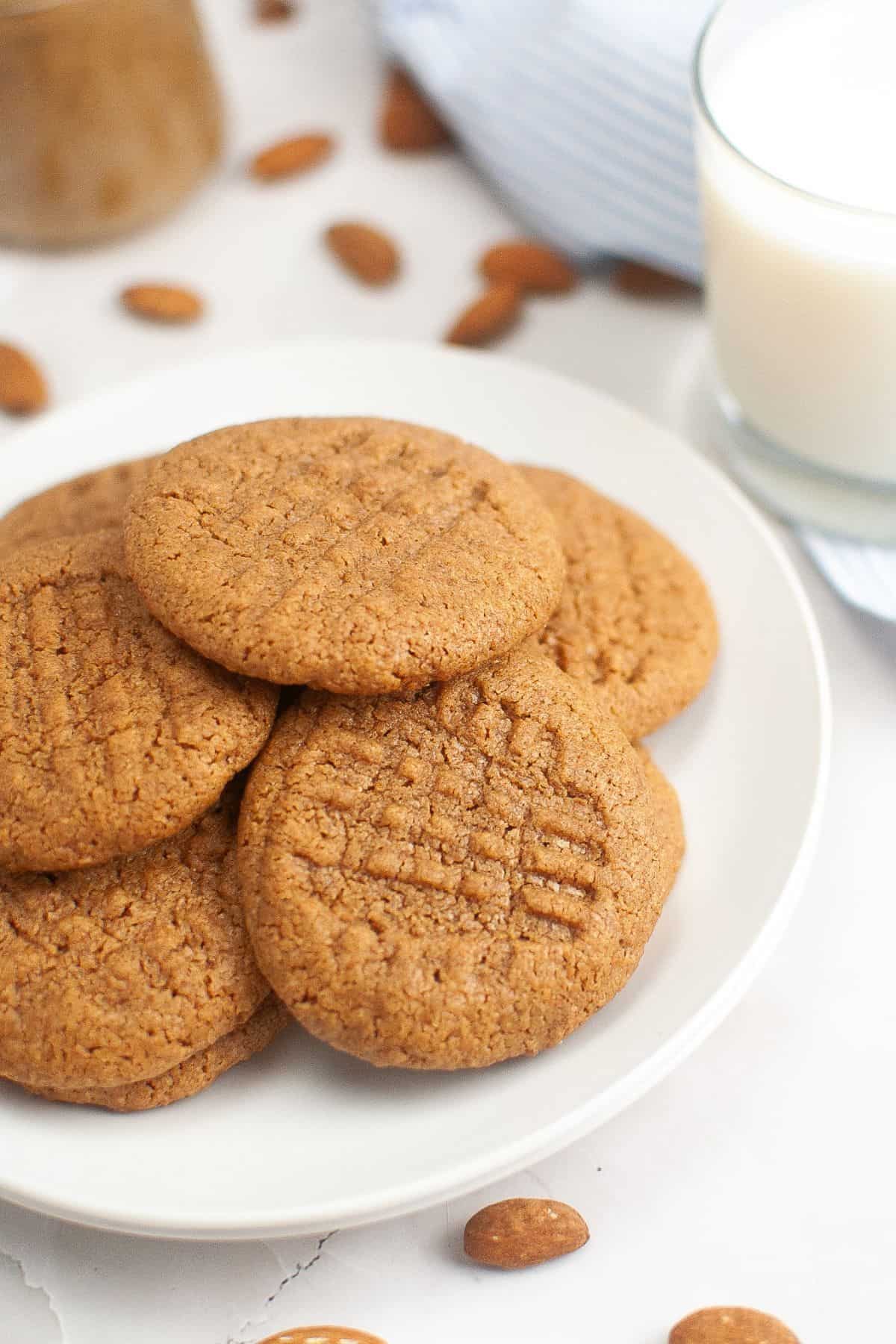 A plate of peanut butter cookies next to a glass of milk, with almonds scattered around on a white surface.