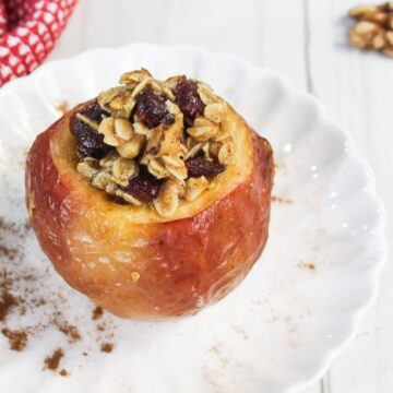 Baked apple in air fryer filled with oats and dried fruit on a white plate, next to a red-checkered cloth and cinnamon sticks.