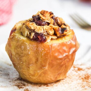 A baked apple in the air fryer brims with nuts, oats, and cranberries on a white plate, adorned with a sprinkle of cinnamon nearby.