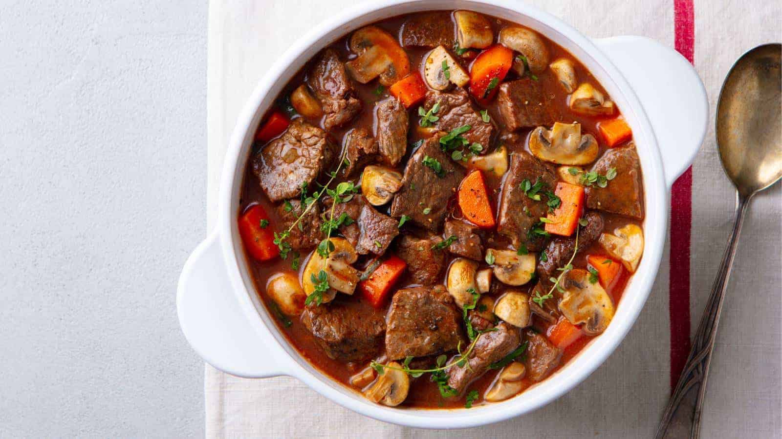 A bowl of beef stew with chunks of meat, sliced mushrooms, carrots, and herbs on a white surface, accompanied by a spoon.