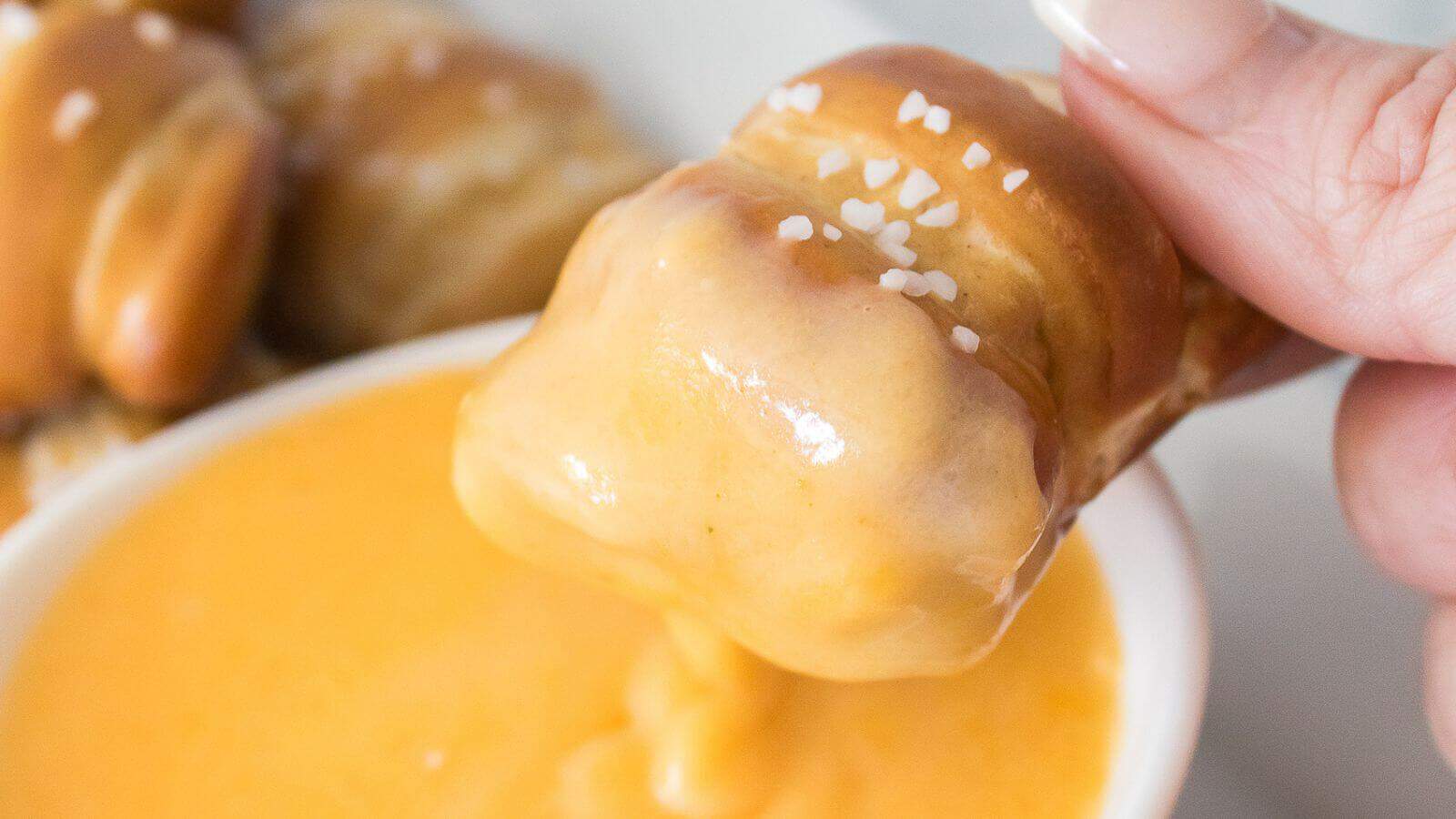 A pretzel bite is being dipped into a bowl of cheese sauce.