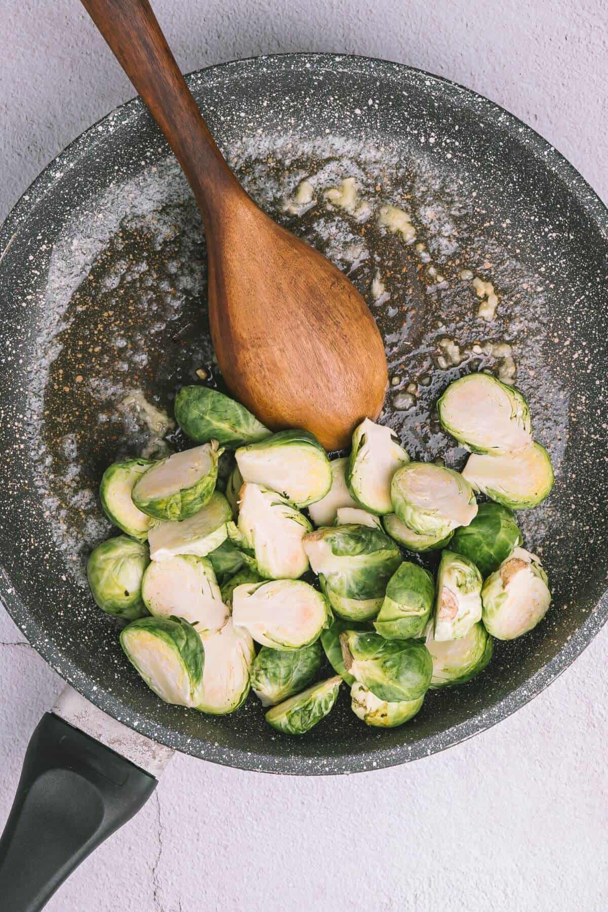 Brussels sprouts being sautéed in a pan with a wooden spoon on a light textured surface.