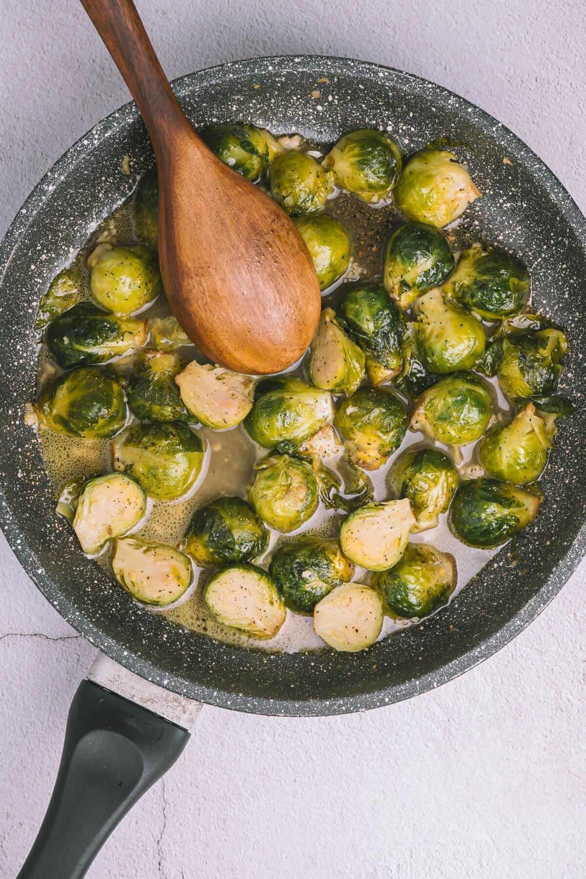 Brussels sprouts cooking in a pan with a wooden spoon on a light-colored surface.