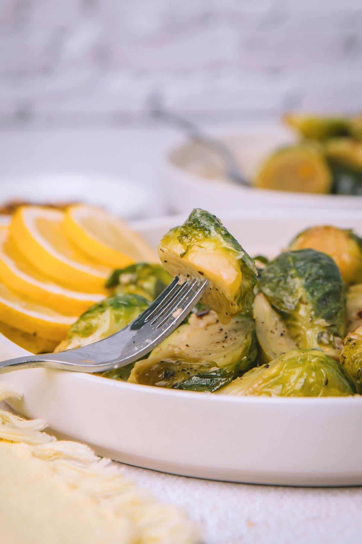 A fork pierces a Brussels sprout in a dish, with lemon slices in the background.