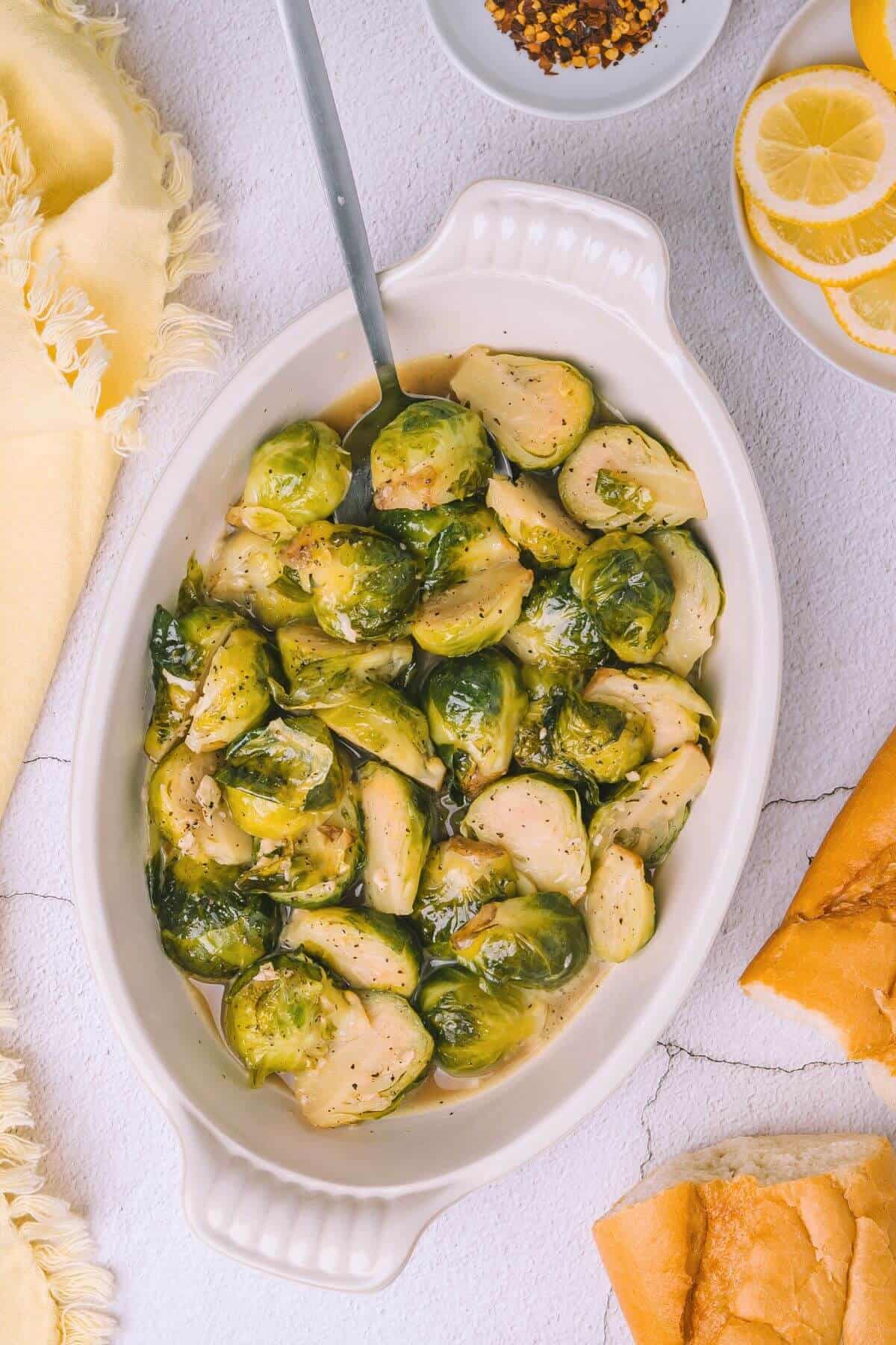 A dish of cooked Brussels sprouts with a spoon on a light textured surface, accompanied by a yellow cloth, red pepper flakes, lemon slices, and bread pieces.