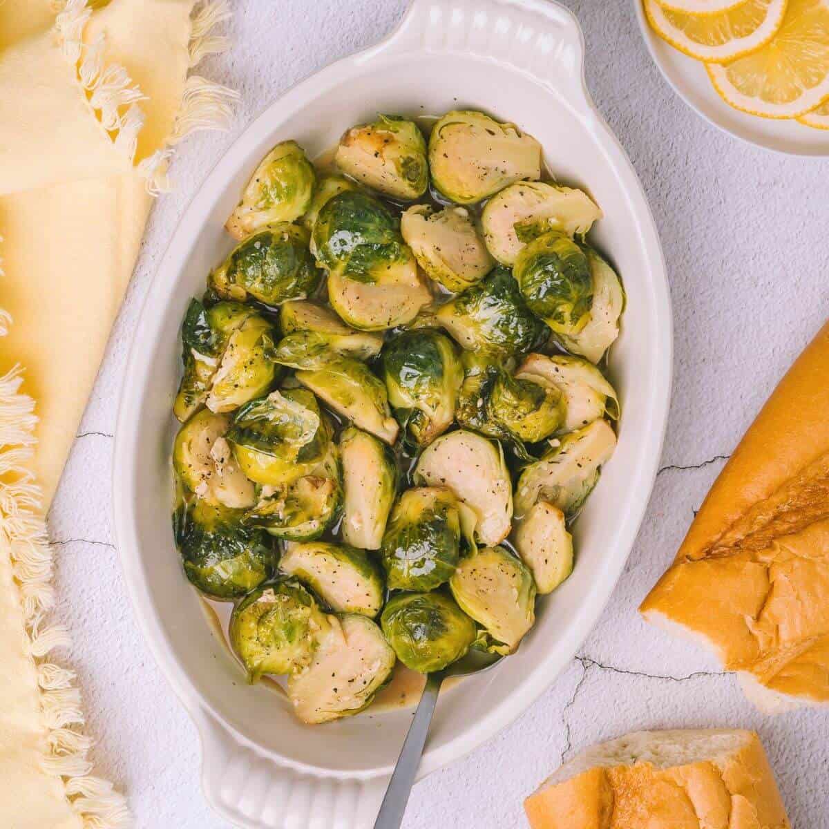 A white oval dish filled with seasoned, roasted Brussels sprouts with a serving spoon. Nearby are lemon slices on a small plate and two pieces of bread on a light-colored surface.
