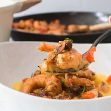 A hand holding a fork with shrimp and vegetables over a bowl of creamy yellow grits, against a kitchen background with a pan and pot.