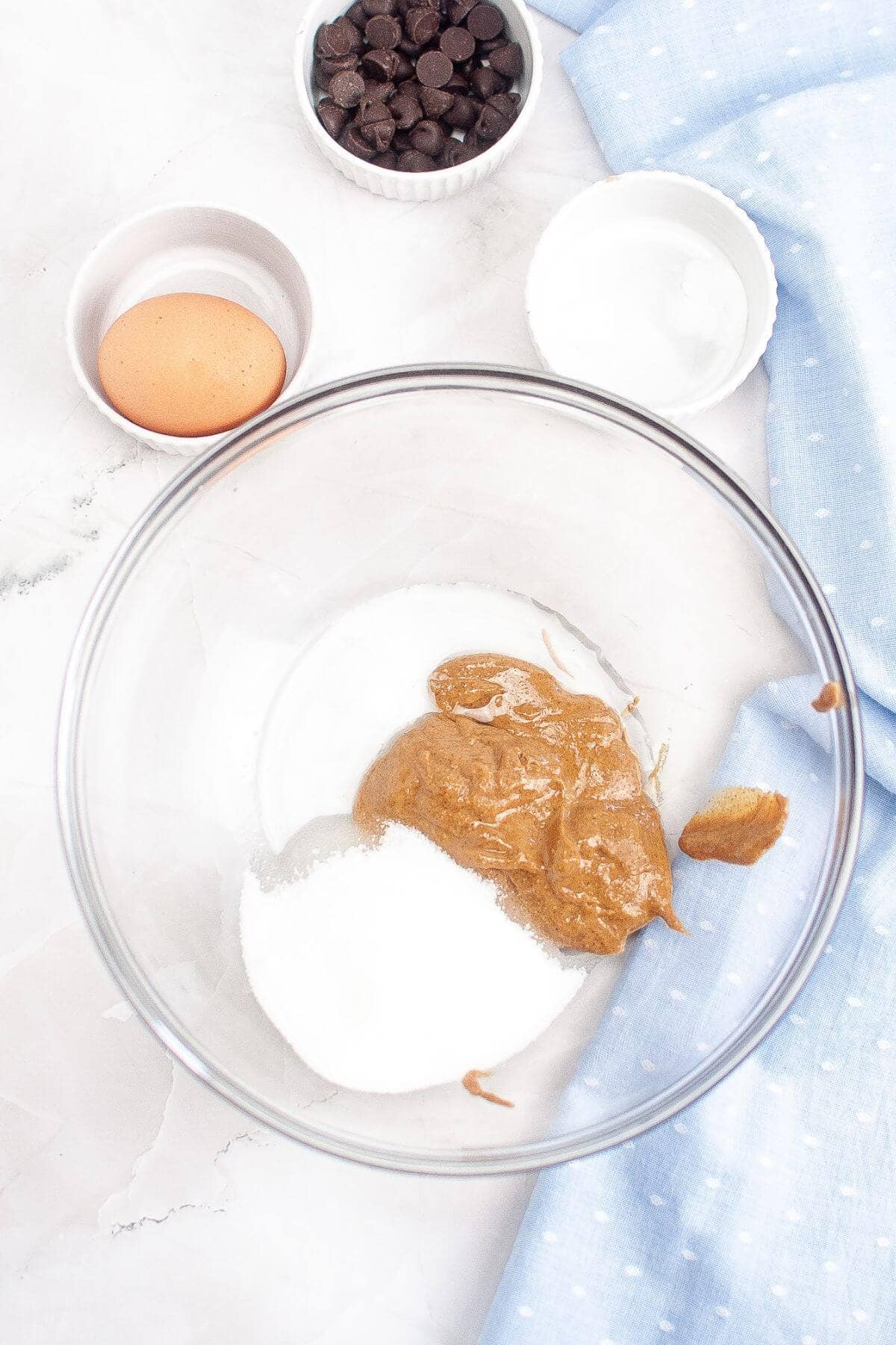 A mixing bowl with peanut butter, sugar, and salt. An egg and chocolate chips are in separate bowls nearby, with a light blue cloth on the side.