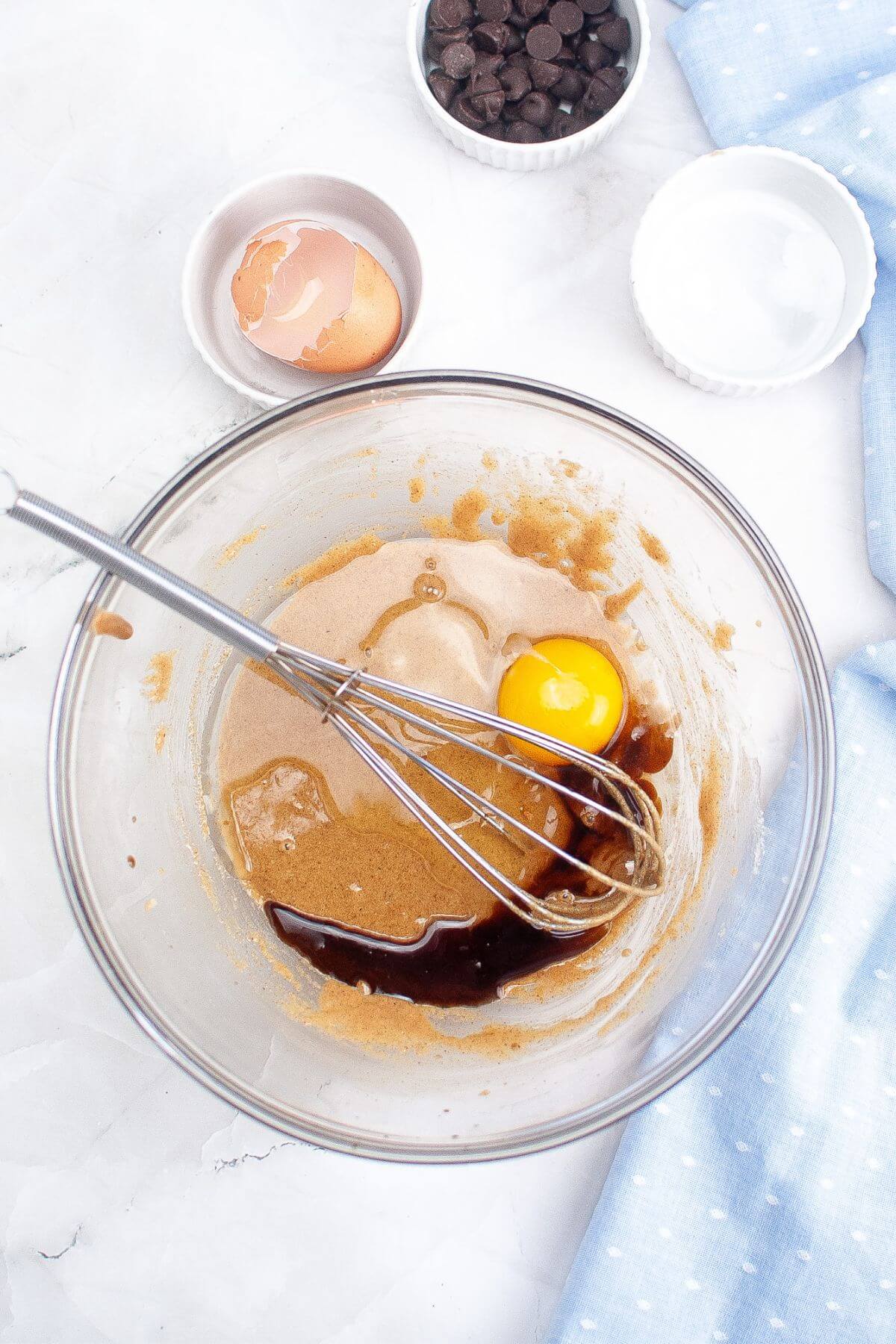 A mixing bowl with batter, a cracked egg, and a whisk. Nearby are a bowl of chocolate chips, an empty bowl with an eggshell, and a bowl containing two white ingredients.