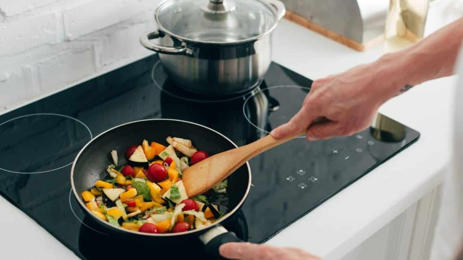 Person stirring a colorful vegetable mix in a frying pan on a stovetop, with a pot in the background.