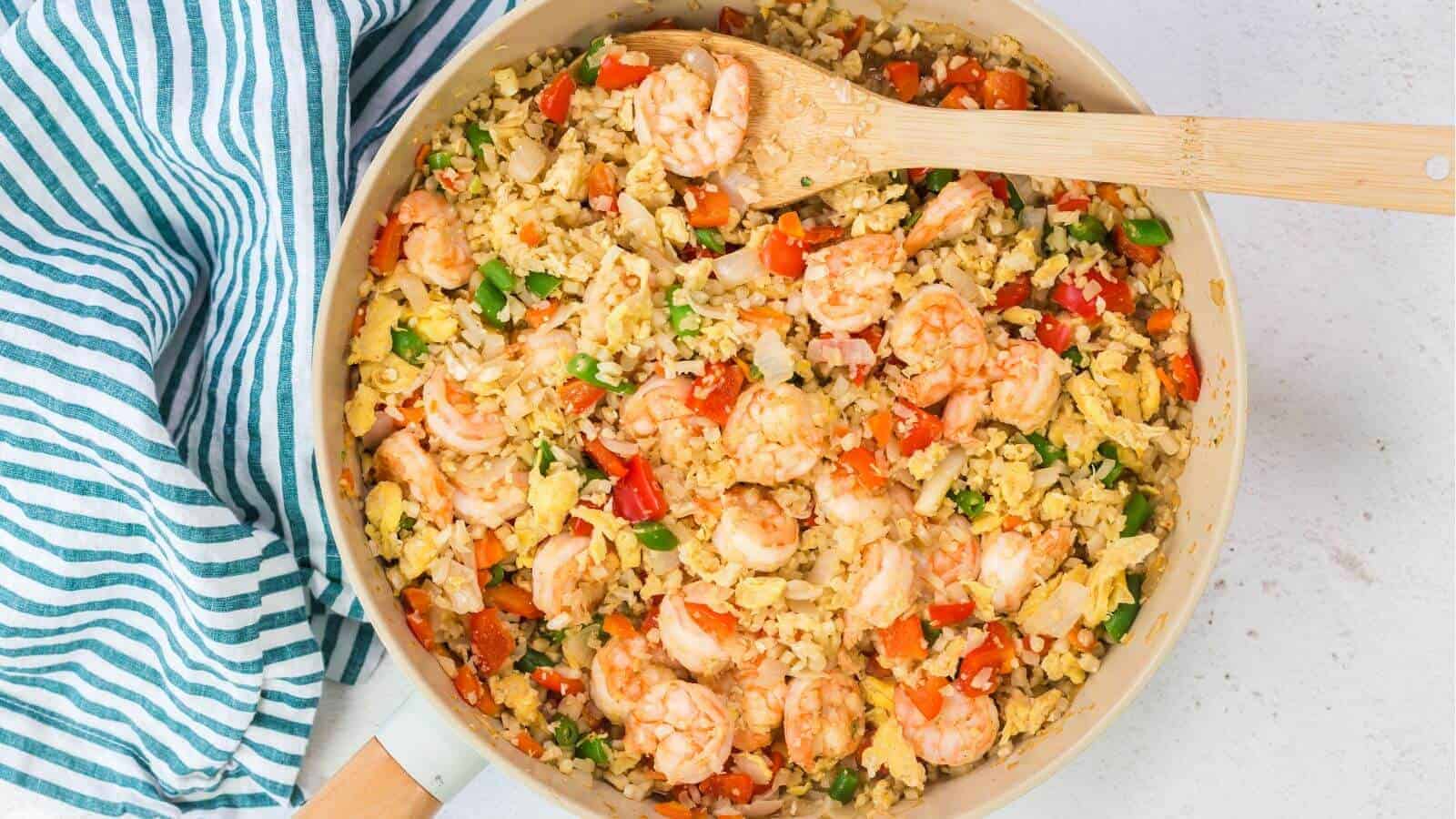 A skillet with shrimp fried rice mixed with vegetables and scrambled eggs, next to a striped cloth.