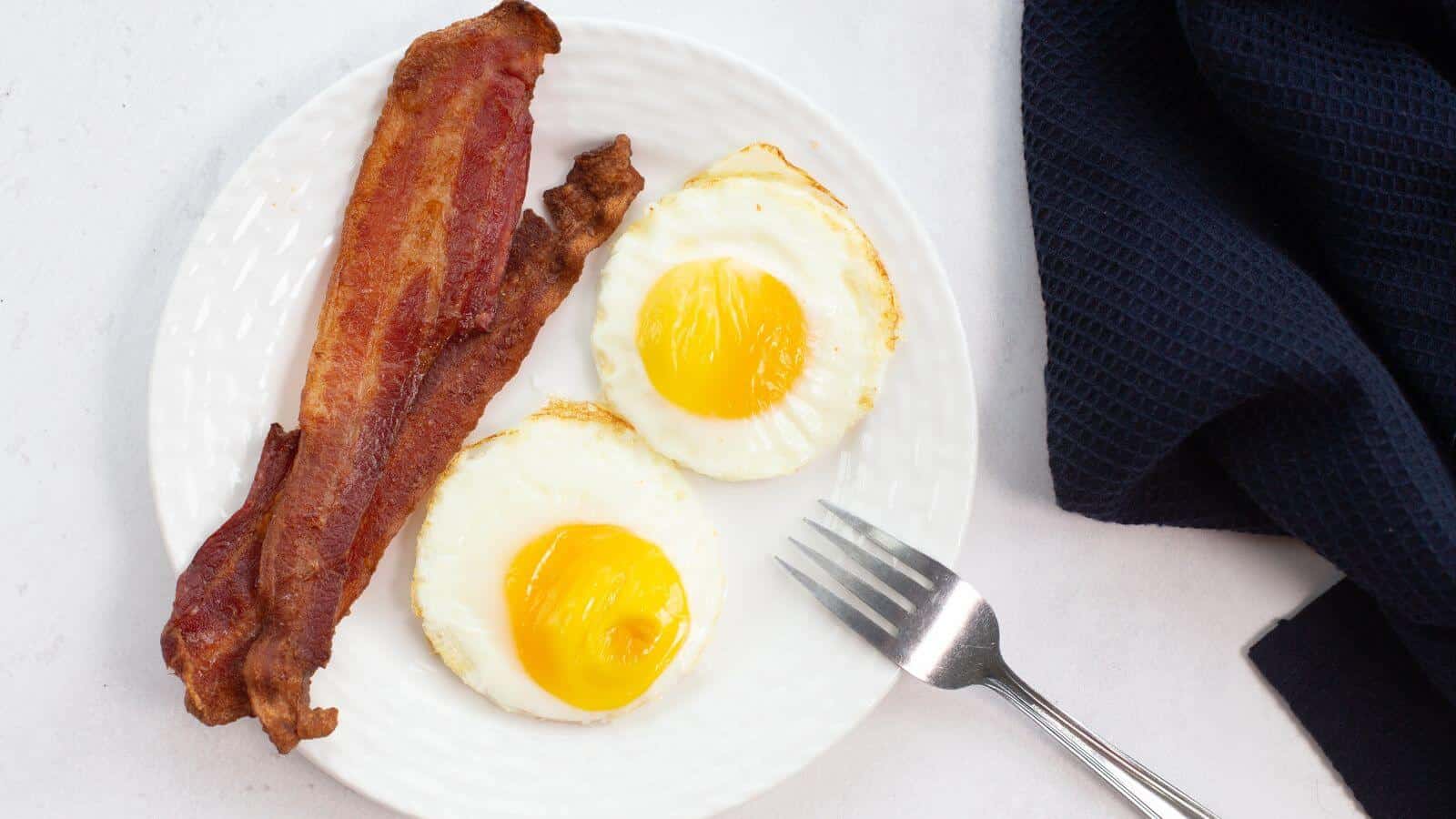 Plate with two sunny-side-up eggs, perfectly cooked like fried eggs in an air fryer, and two strips of crispy bacon, next to a fork and a dark cloth napkin.