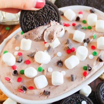 A hand dips a chocolate sandwich cookie into a cream cheese dip topped with mini marshmallows, chocolate chips, and colorful sprinkles. Surrounded by more cookies and graham crackers.