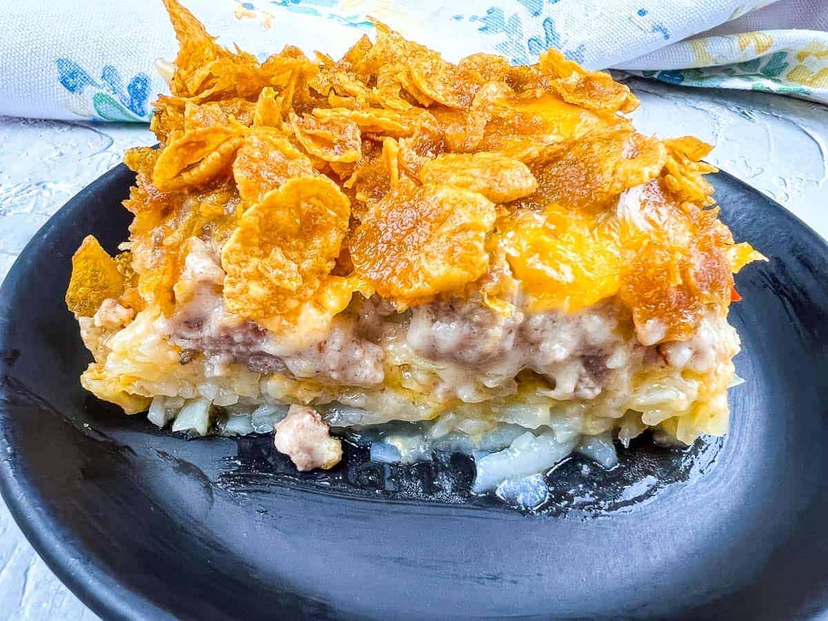 A close-up of a casserole slice on a black plate, showcasing a crispy cornflake topping over layers of cheese, meat, and potatoes—these casseroles taste like a chef made them but you did.