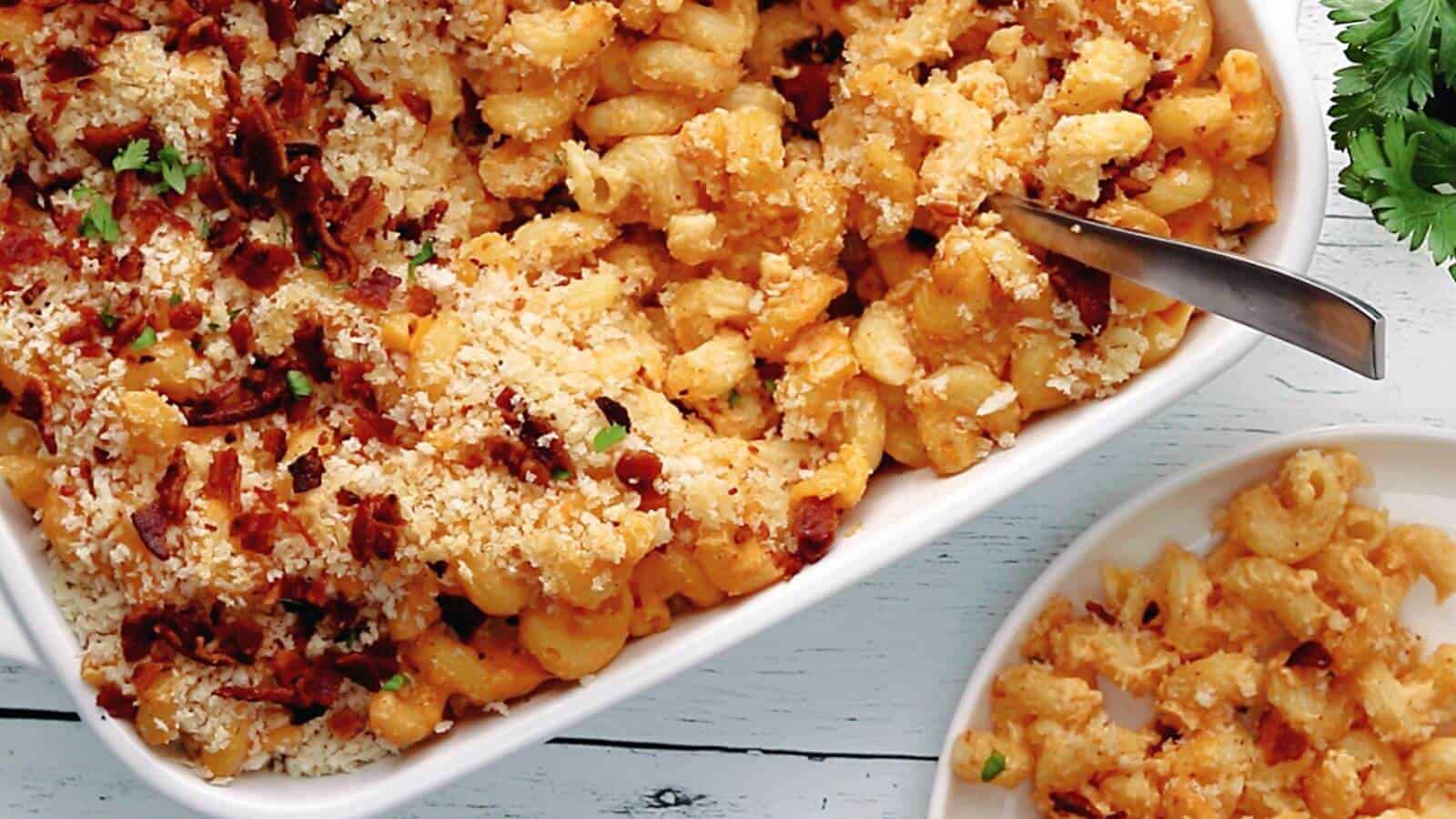 Baked macaroni and cheese topped with breadcrumbs and bacon in a white dish, with a serving on a plate beside it.